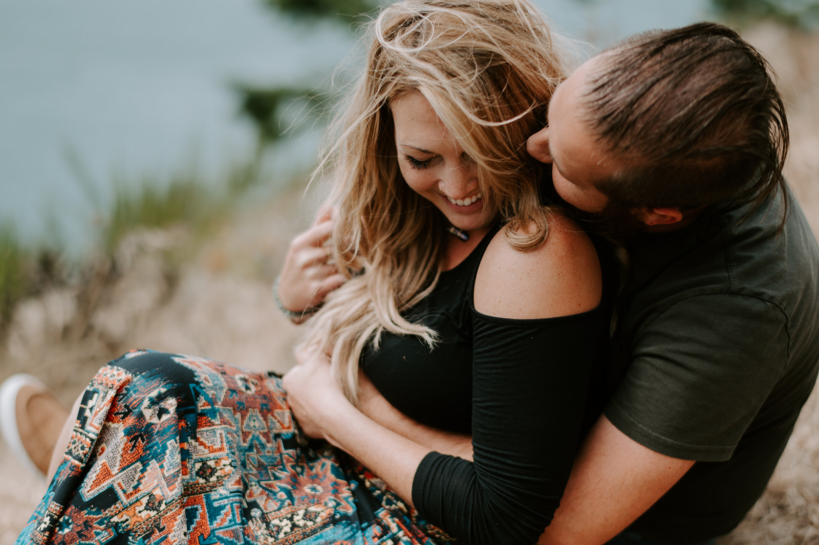 Couple sitting on a grassy hill, embracing and smiling with a serene view of the ocean in the background.