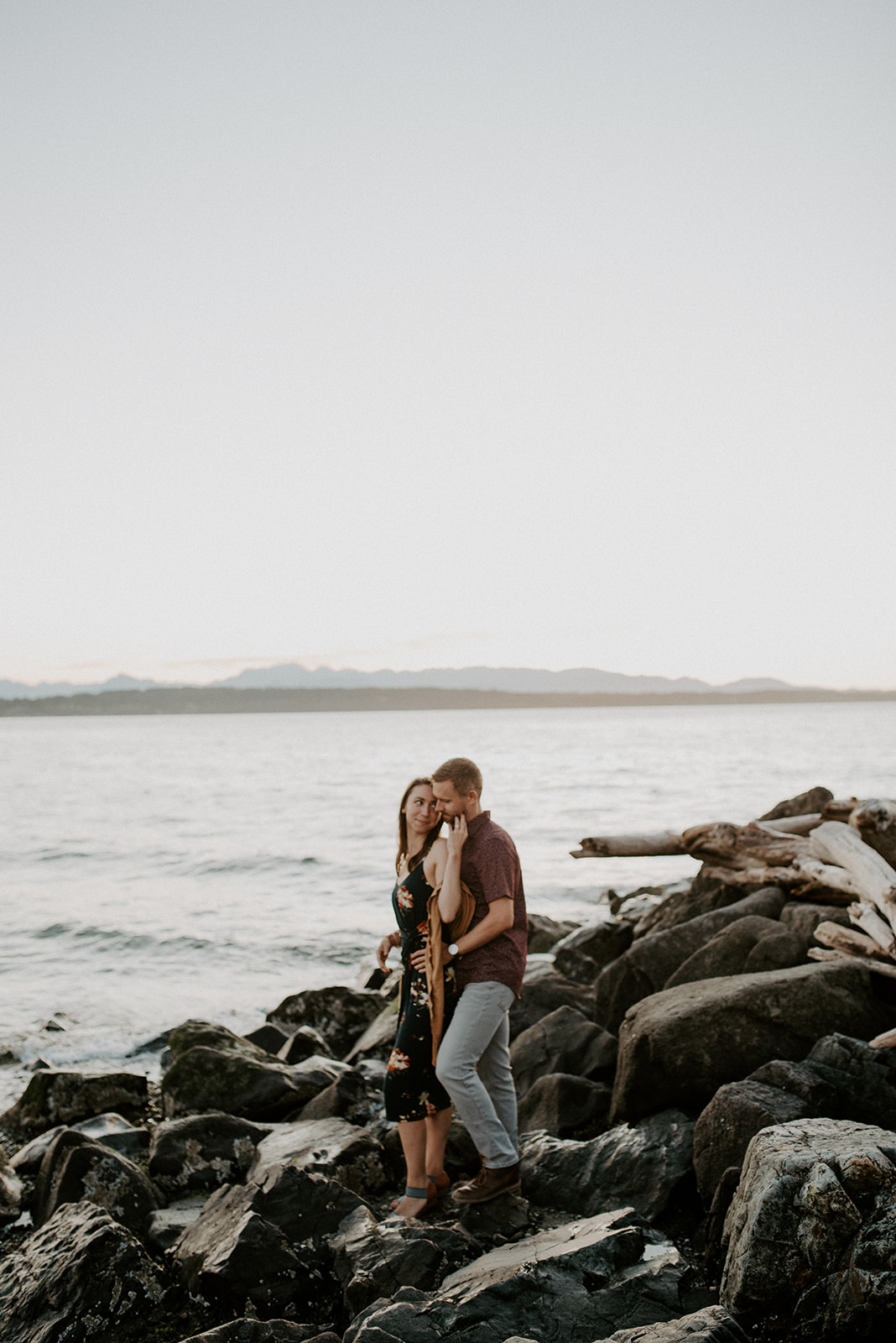 Couple standing on rocky shores at Discovery Park with Puget Sound and distant mountains in the background.