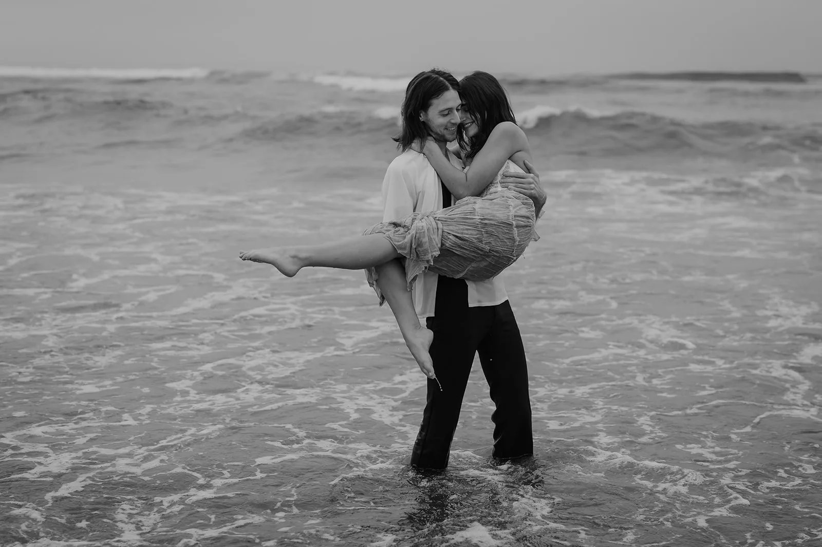 Swept Away - Romantic Moment on the Oregon Coast Man carrying woman in his arms on the beach in a black and white Oregon coast engagement photo