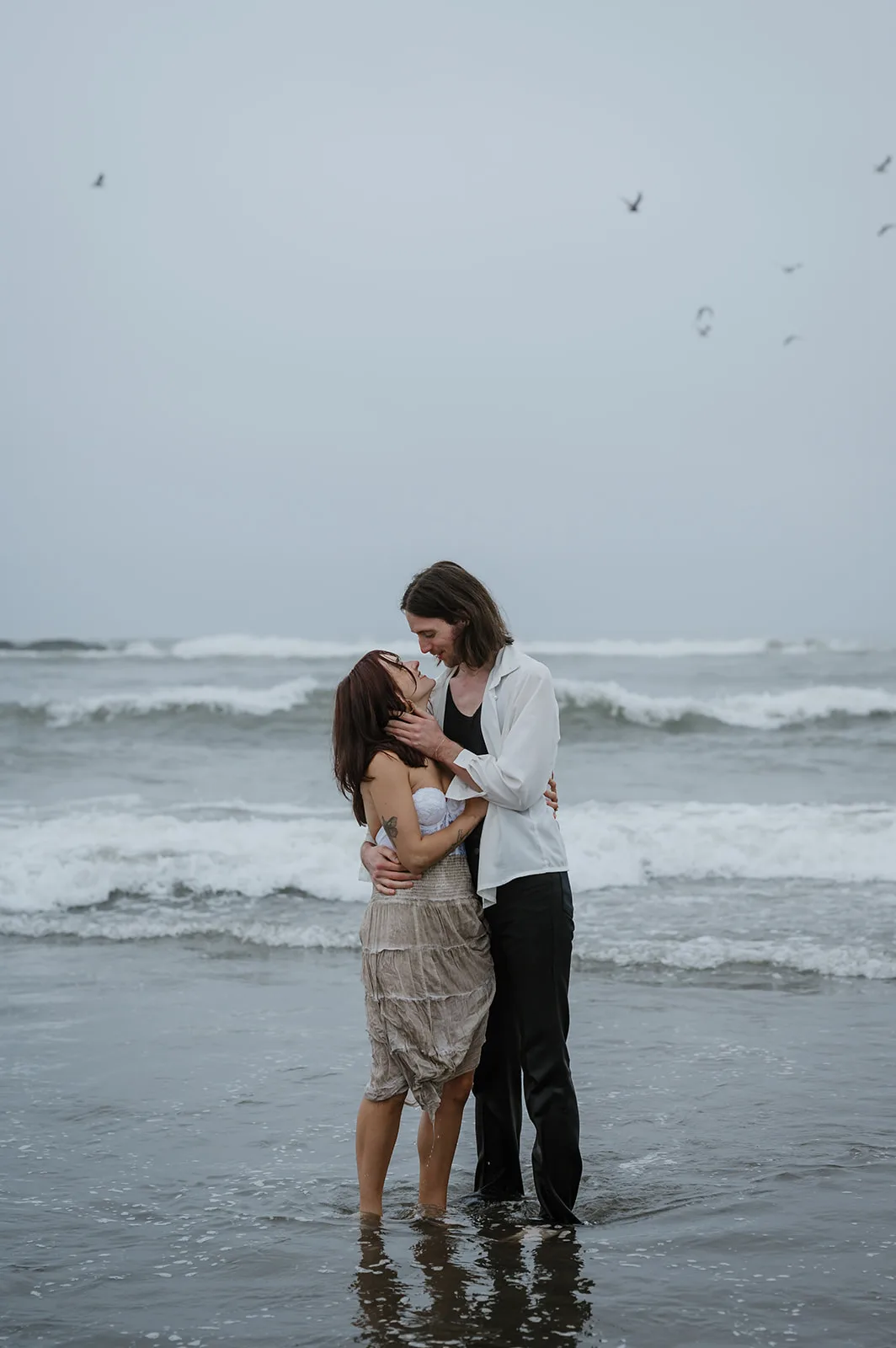Ocean's Embrace - Tender Engagement on the Oregon Shore Couple sharing a tender moment amidst the ocean waves on a cloudy day