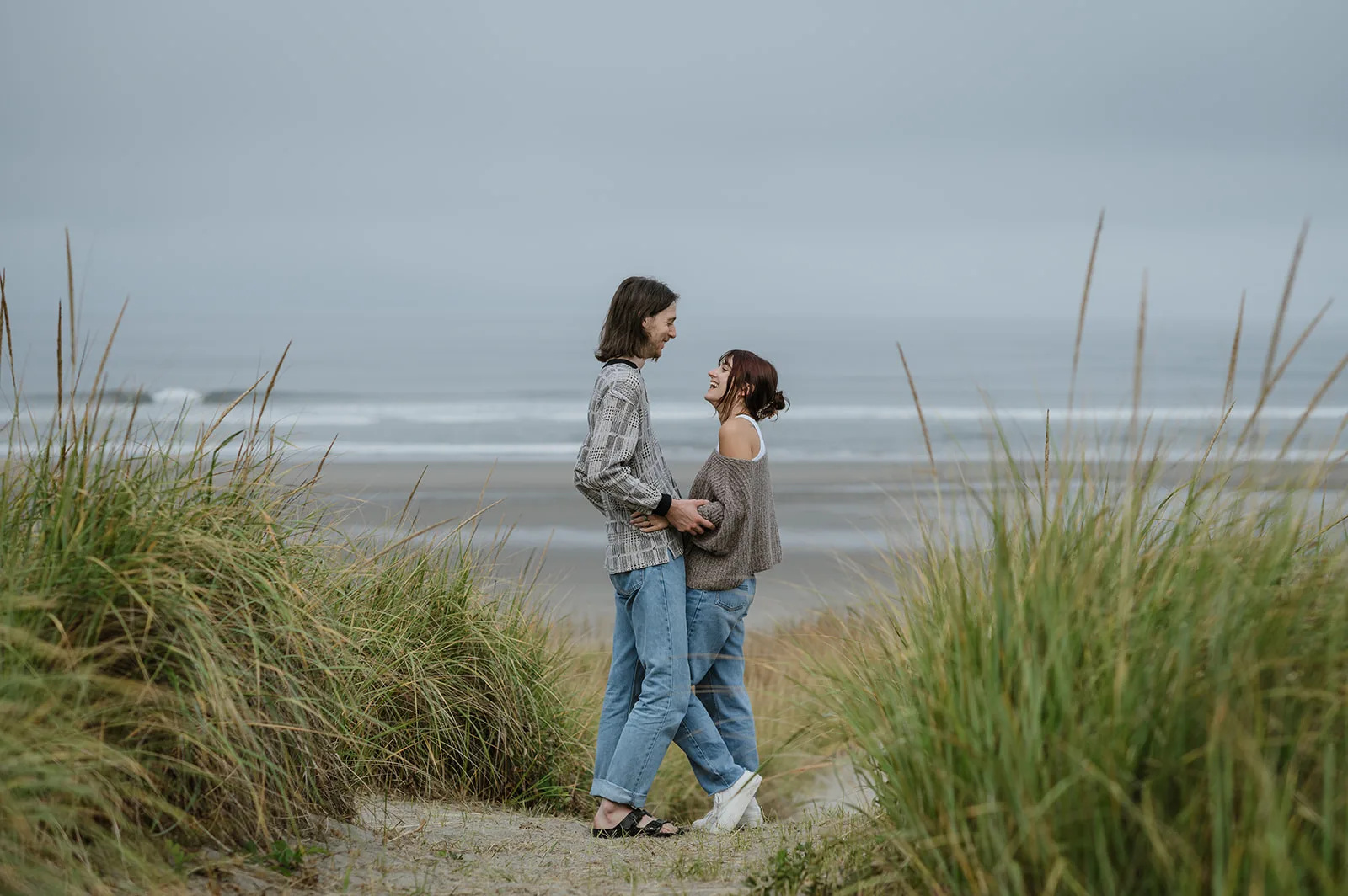 Seaside Romance - Oregon Coast Beach Engagement Photo Couple standing by the beach grass with ocean waves in the background on the Oregon coast