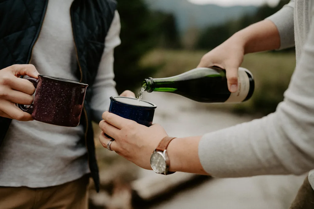 "A close-up of hands pouring champagne into enamel mugs during an engagement celebration in nature."