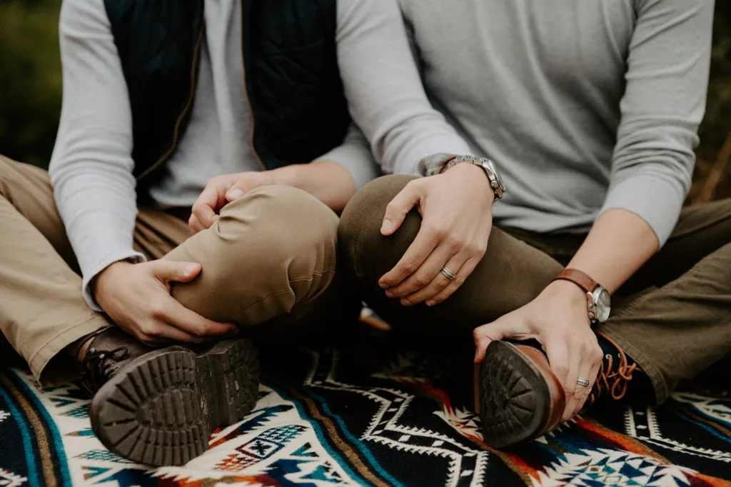"Close-up of a same-sex couple's hands gently touching as they sit closely together on a vibrant, patterned blanket outdoors."