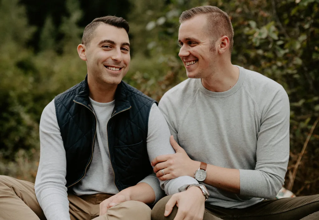 "Two men sitting close, sharing a joyful moment with genuine smiles amidst a natural backdrop."