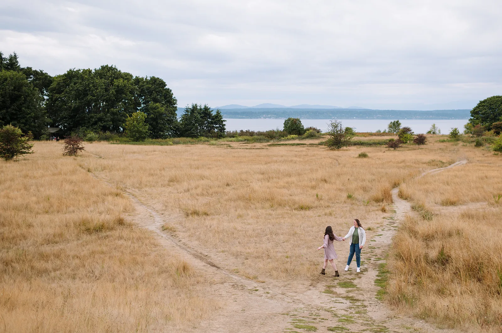 Couple walking hand-in-hand through a wide open field at Discovery Park with Puget Sound in the background.