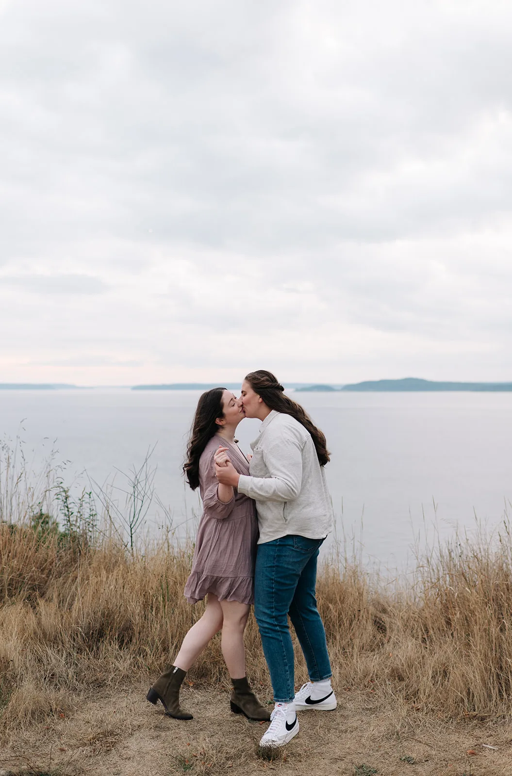 Couple sharing a kiss on a grassy hill at Discovery Park with a scenic water view behind them.