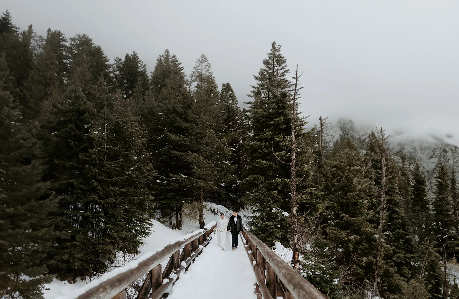 Bride and groom walk hand in hand across a snow-covered bridge.