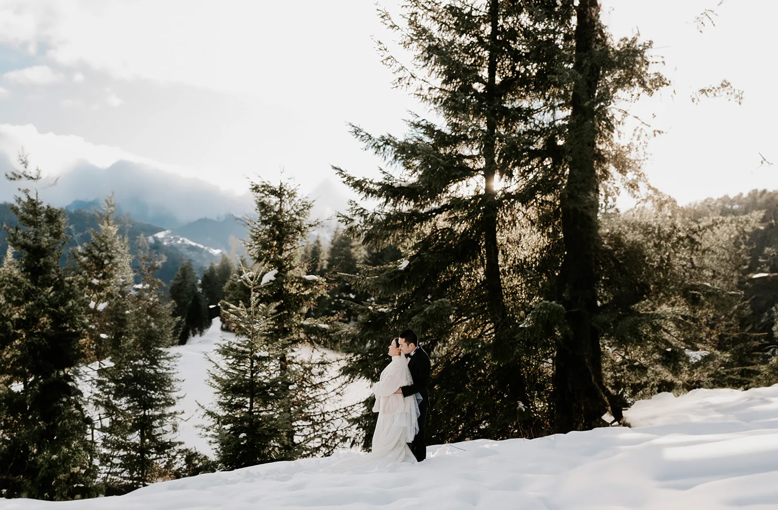 A couple shares an intimate cuddle in the snow-covered pine forest near Gold Creek Pond, with the sun casting a warm glow on their snowy mountain elopement.