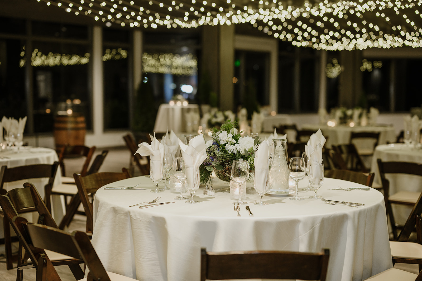 Round table set with white tablecloth, folded napkins, and floral centerpiece under string lights at Dockside at Duke's.
