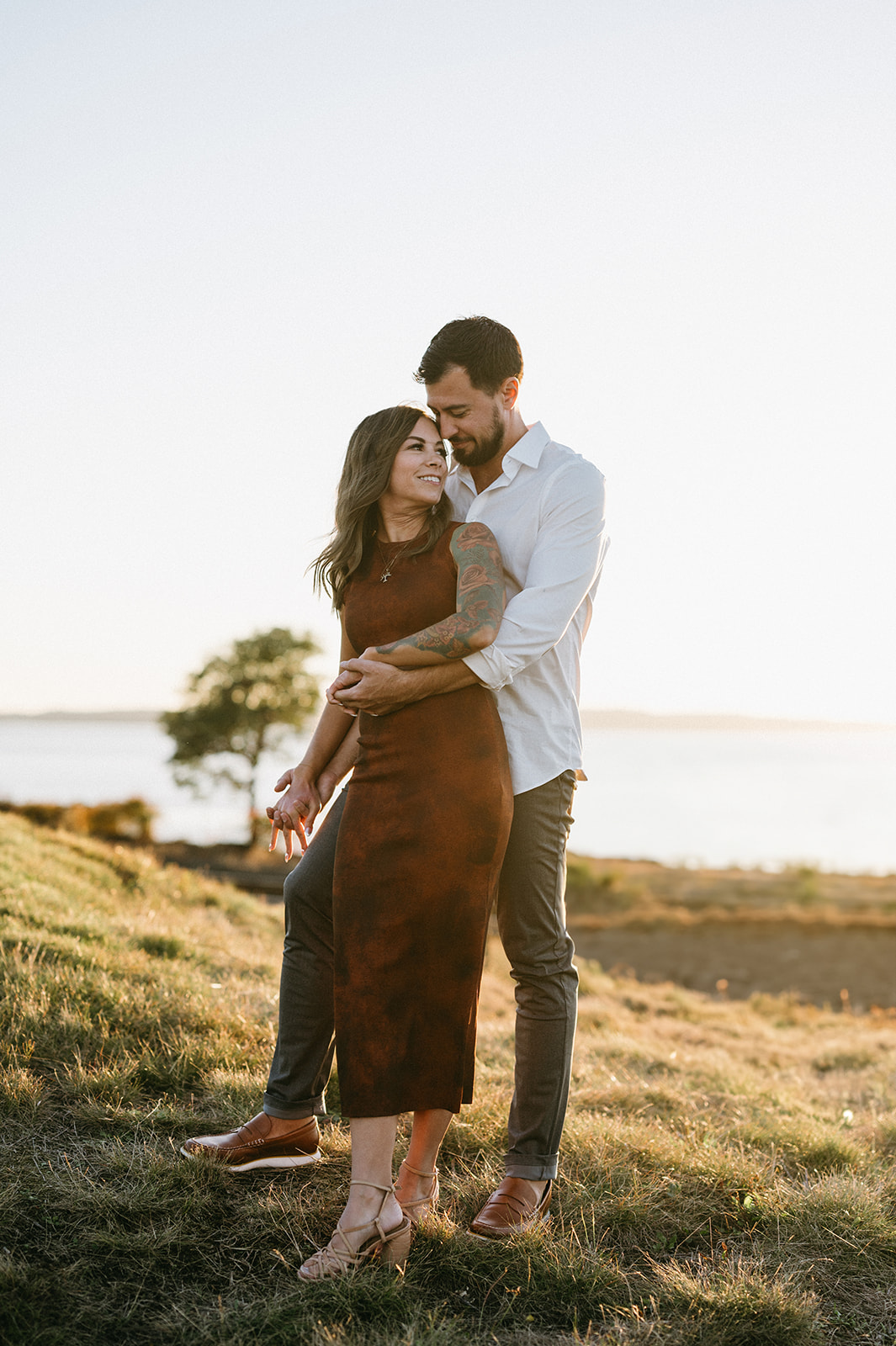 A couple standing on a grassy hill, the man embracing the woman from behind.