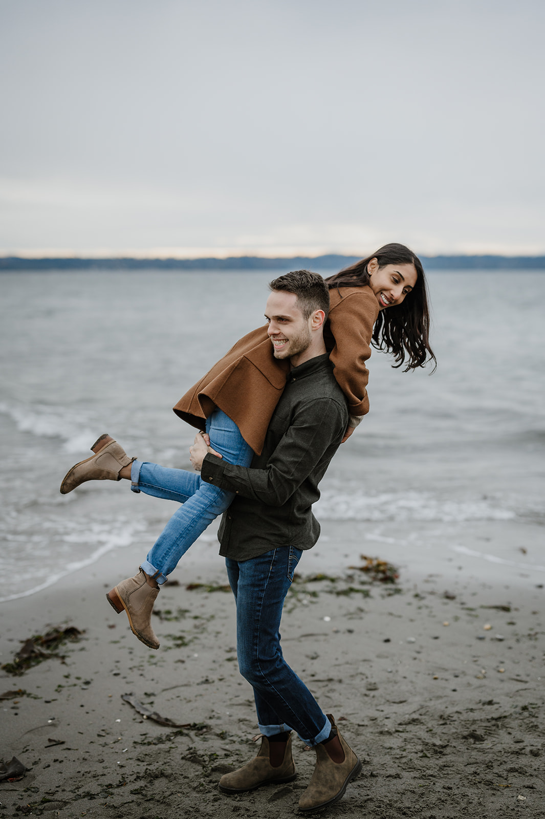 Playful couple on the beach at Discovery Park, with the man lifting the woman as they laugh together.
