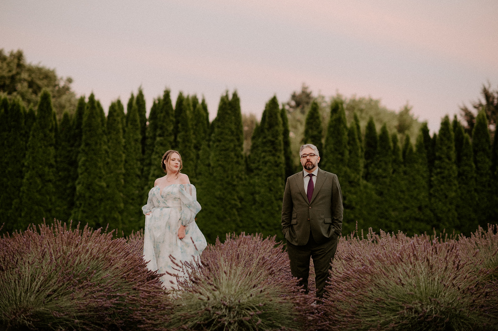 Couple standing apart among rows of lavender with a soft pink sky