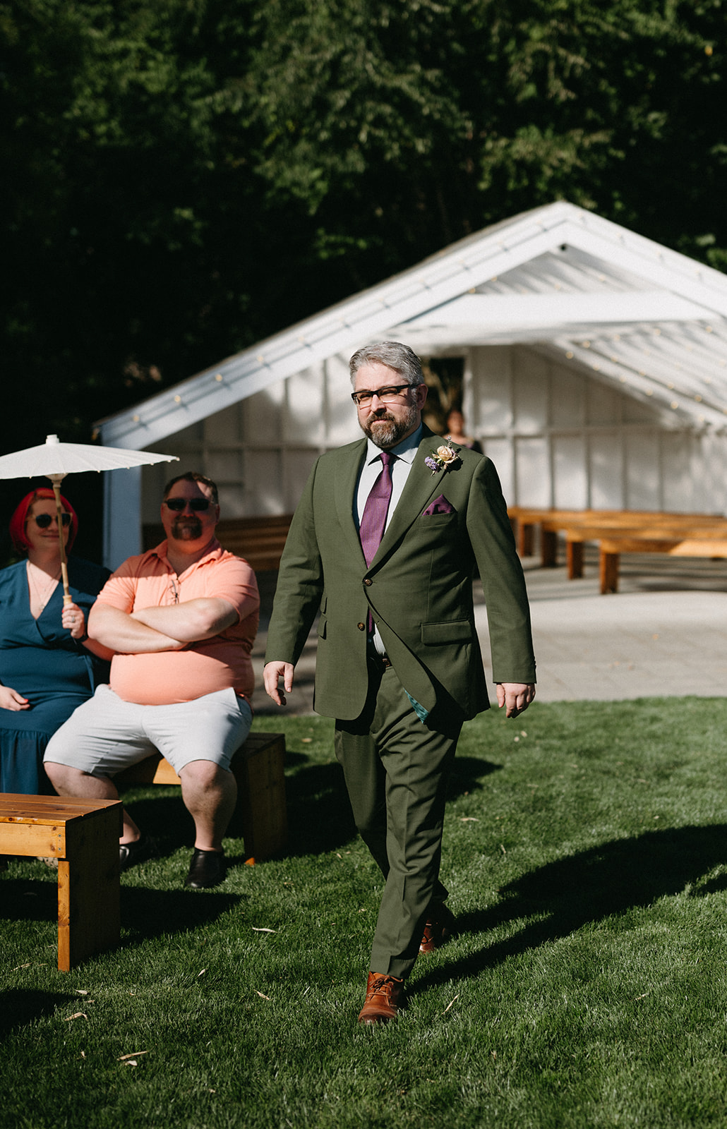 Groom walking down grassy aisle in green suit past seated guests