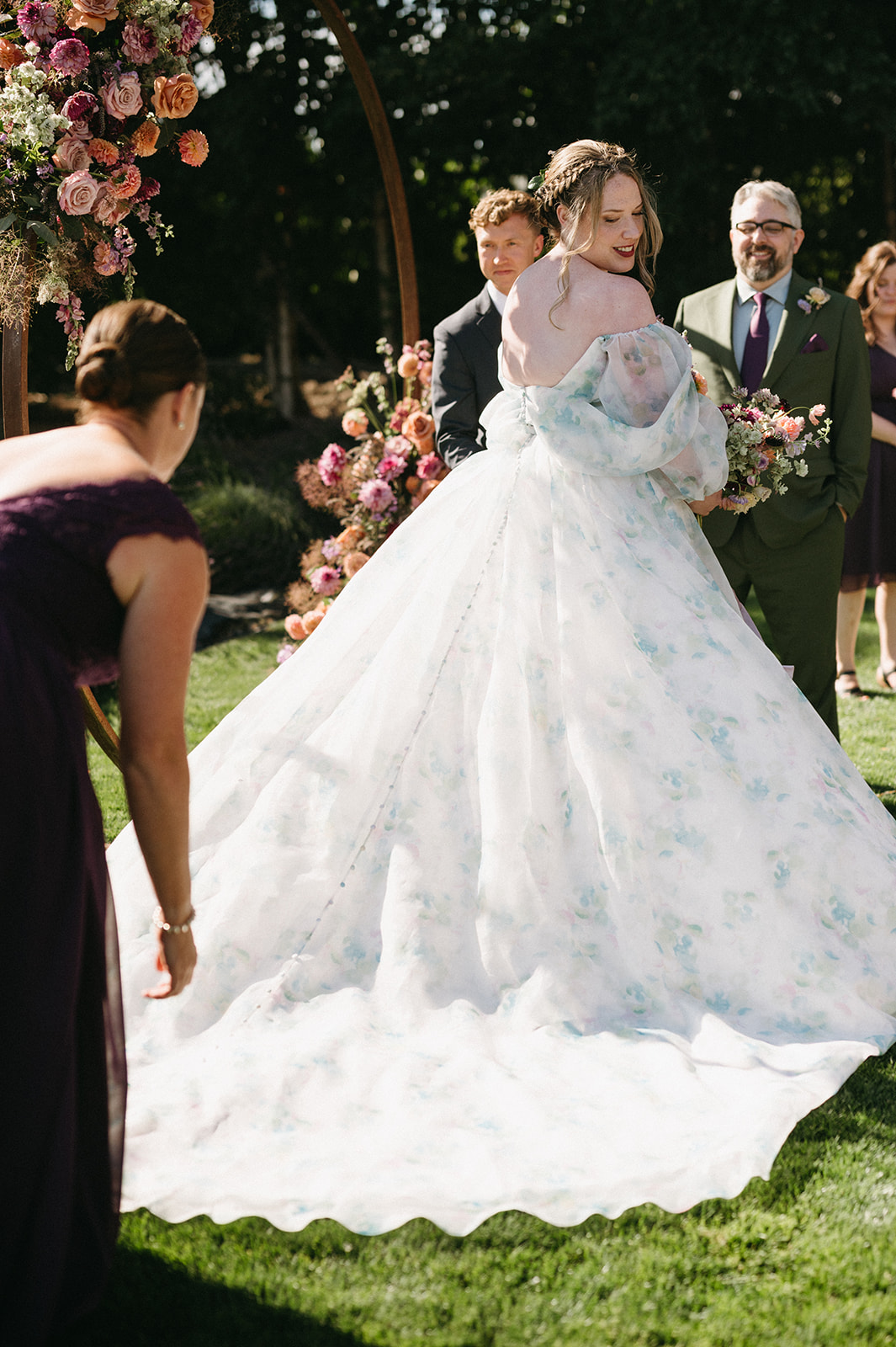 Wedding dress being adjusted as bride stands in sunlight at floral ceremony arch