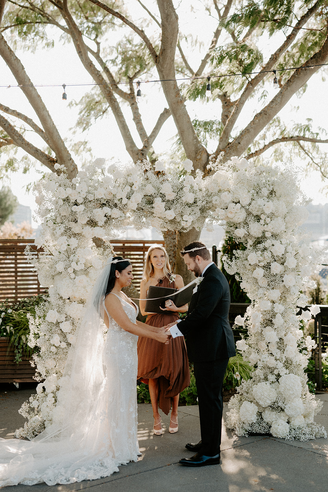 The couple exchanging vows under a large tree, with the officiant in the background during their outdoor wedding.
