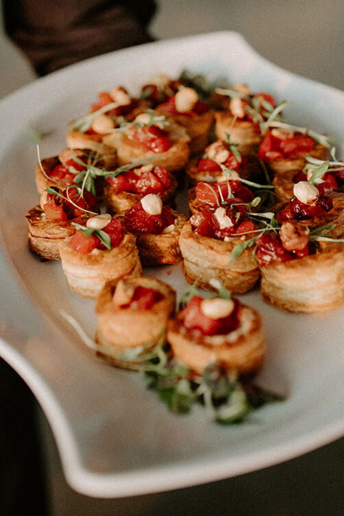 Close-up of a white platter holding bite-sized puff pastry appetizers topped with tomatoes, nuts, and microgreens.