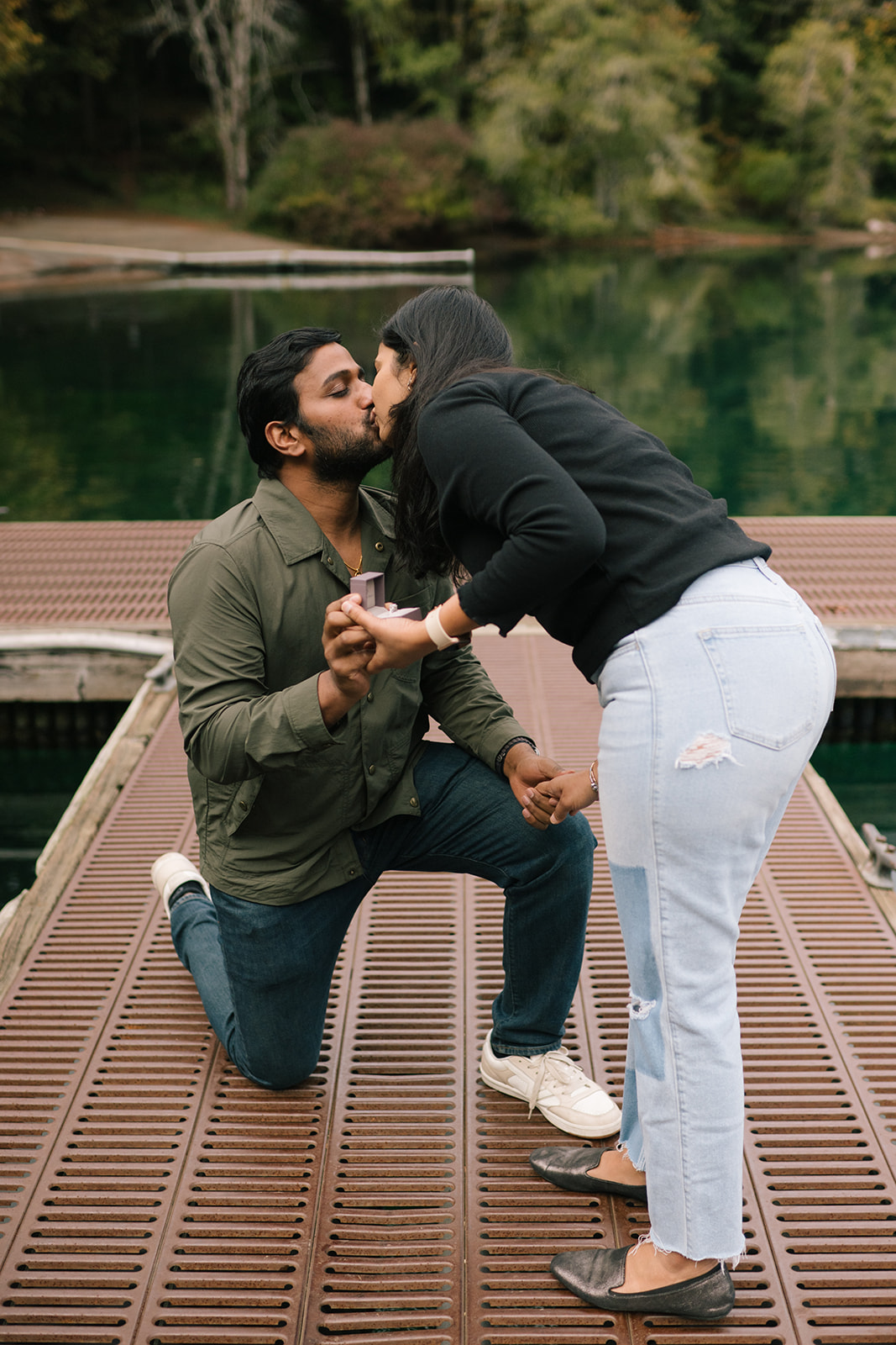 Madhu leaning in to kiss Sudesh as he kneels with the engagement ring at Lake Crescent.