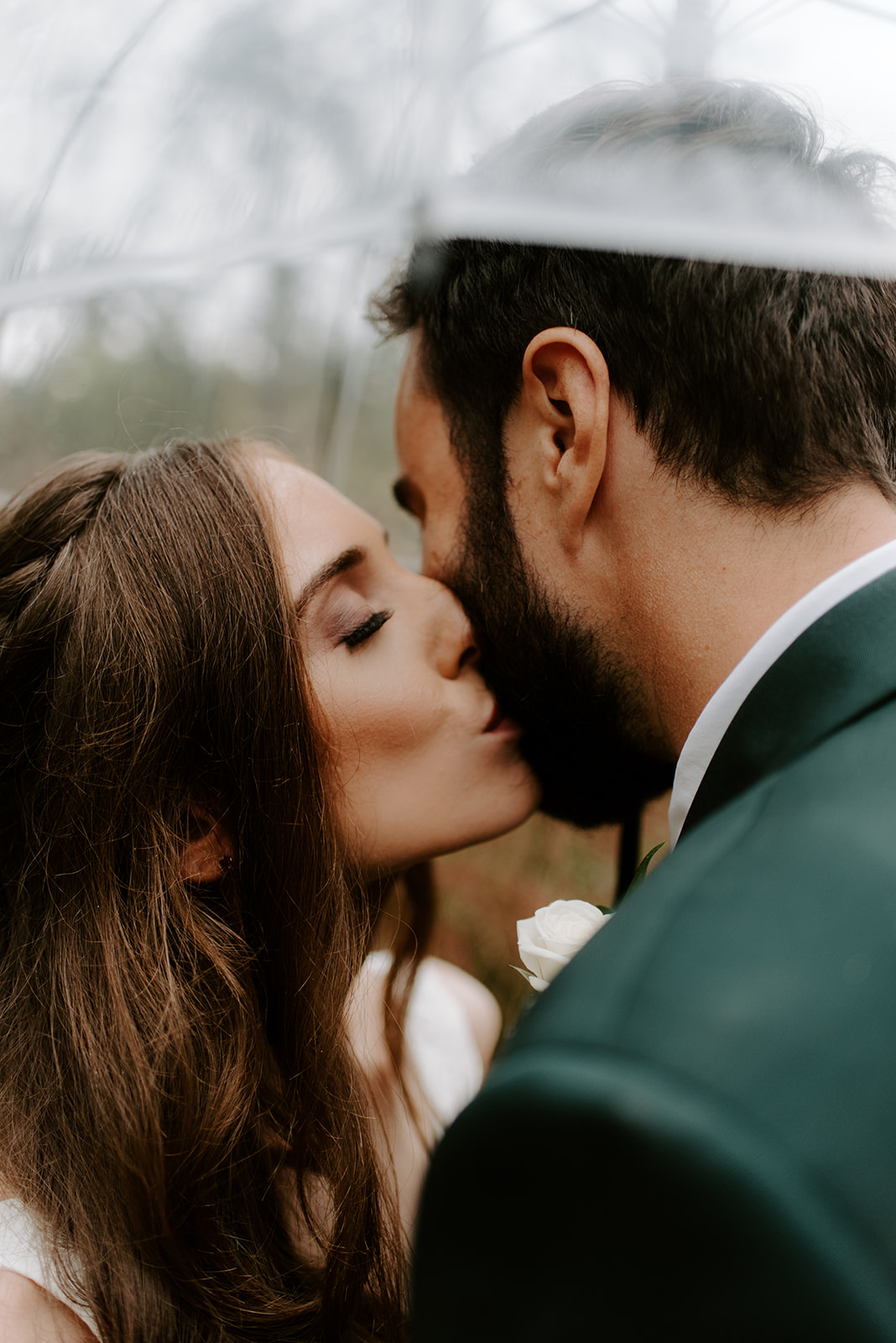 Close-up of a bride and groom sharing a kiss under a clear umbrella on a rainy day.