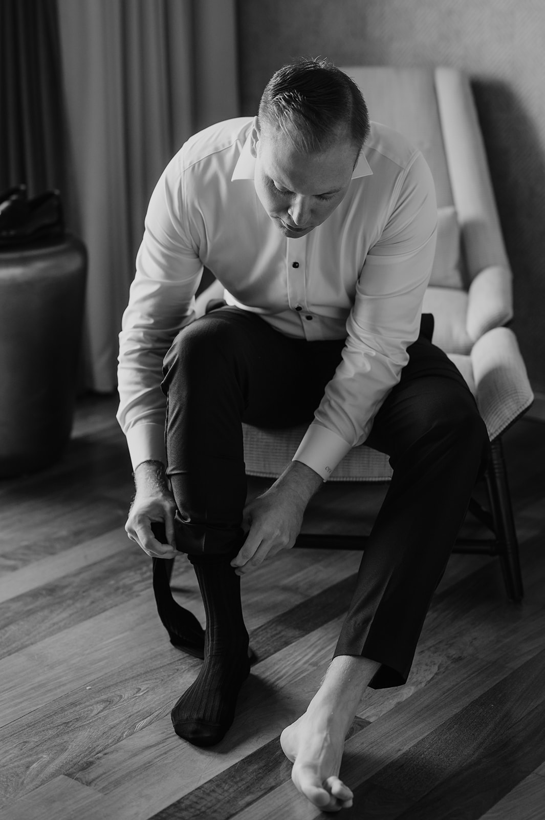 Black and white photo of the groom sitting on a chair pulling on his socks while getting dressed for his wedding day in Maui.
