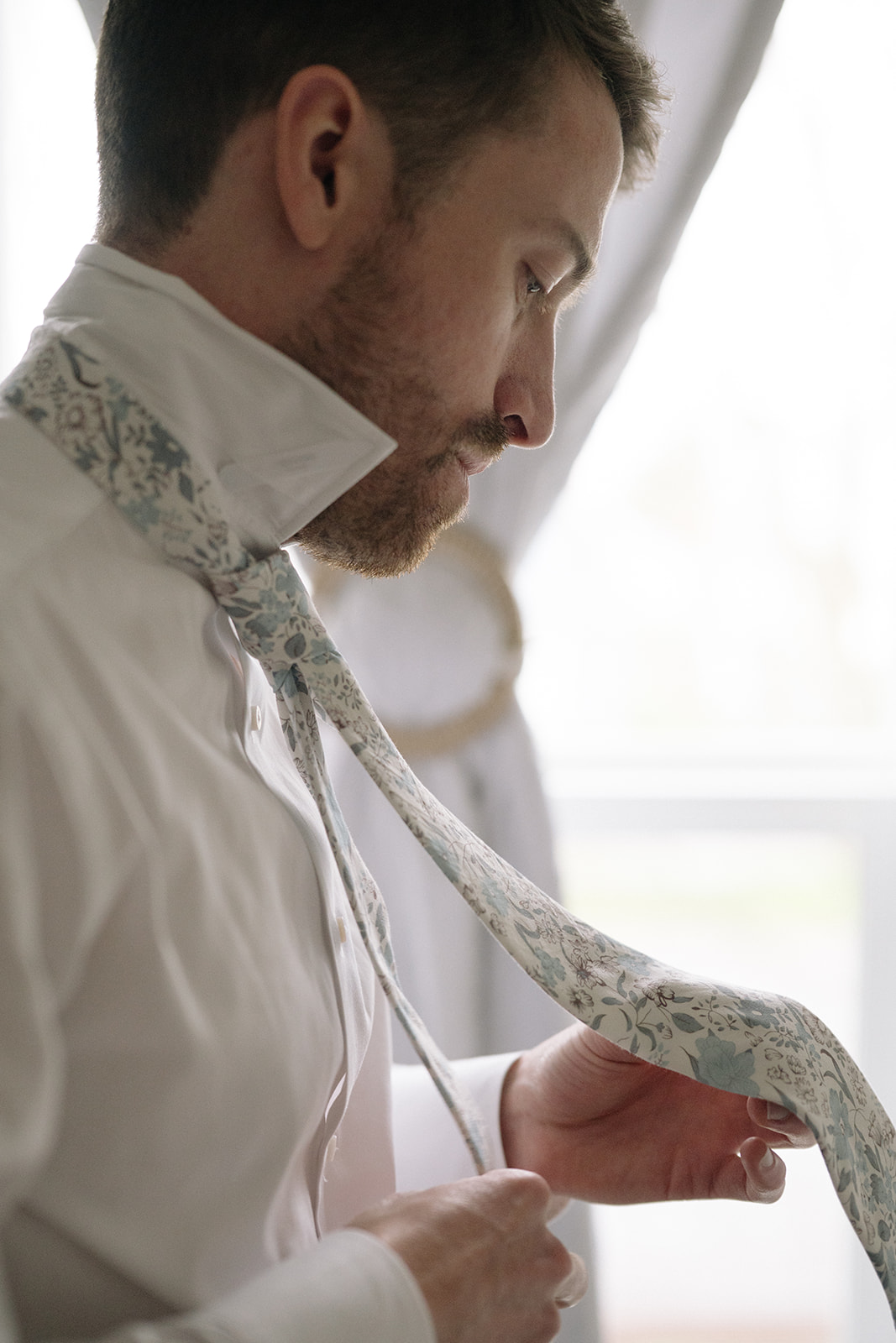 A groom carefully tying a patterned tie in soft window light, focused and serene.