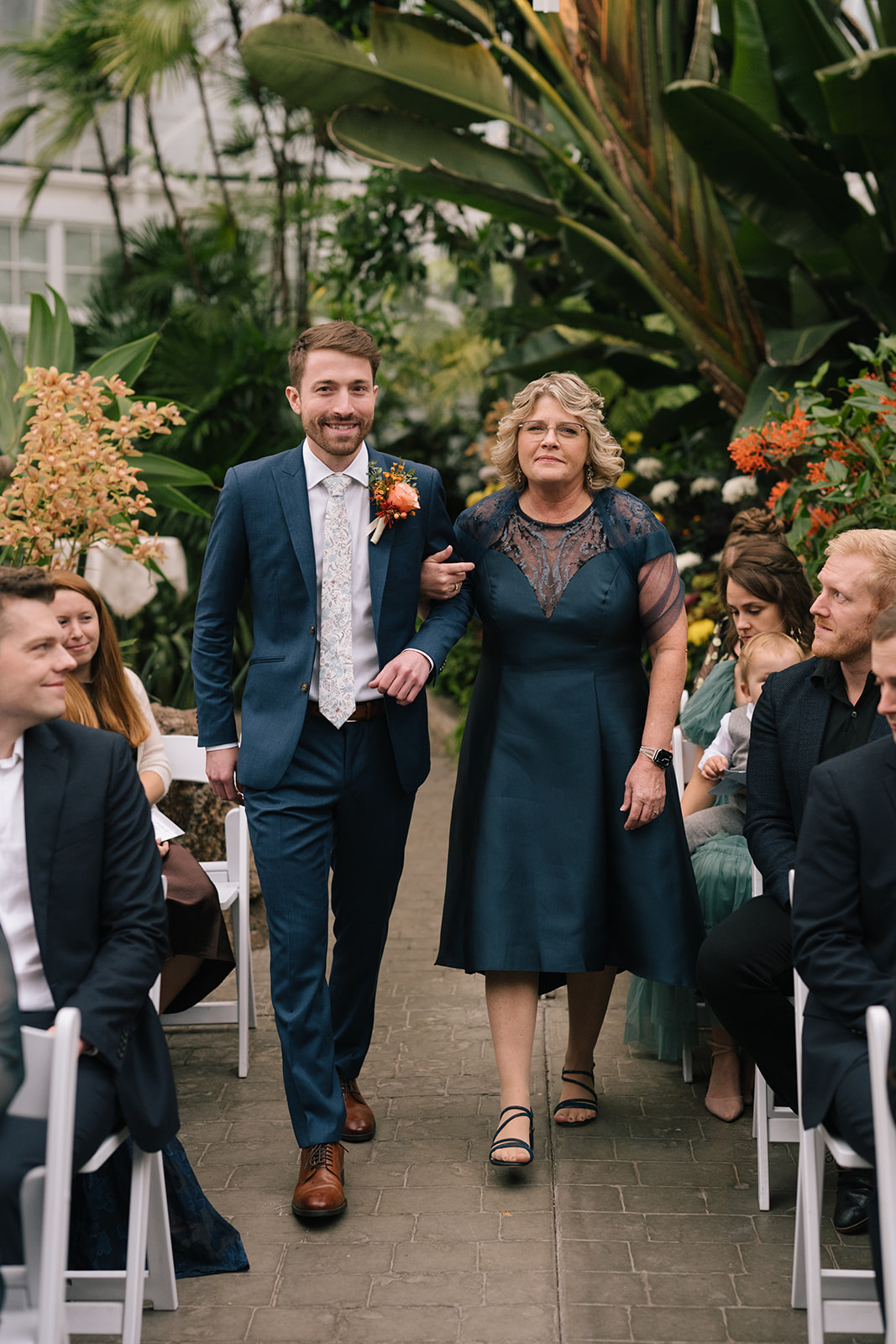 Patrick walking his mother down the aisle surrounded by vibrant greenery and blooms.
