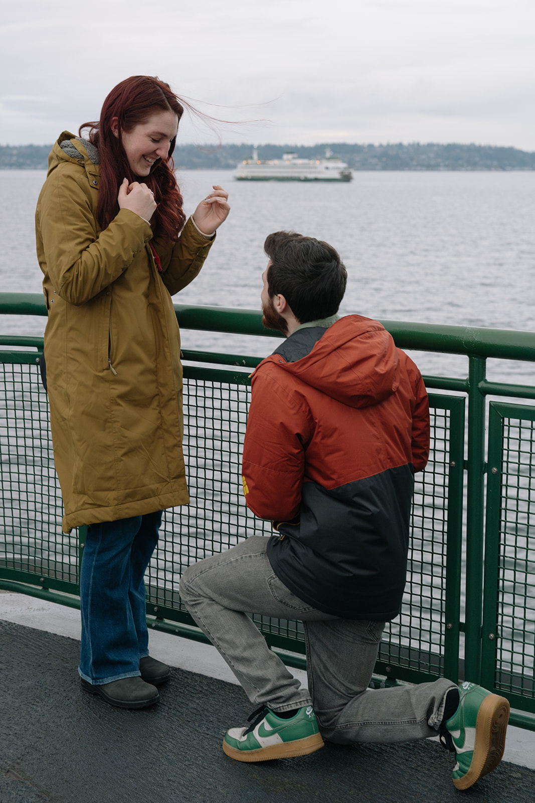 A man kneels to propose on a Washington State ferry as his partner smiles, with water and a distant ferry in the background.