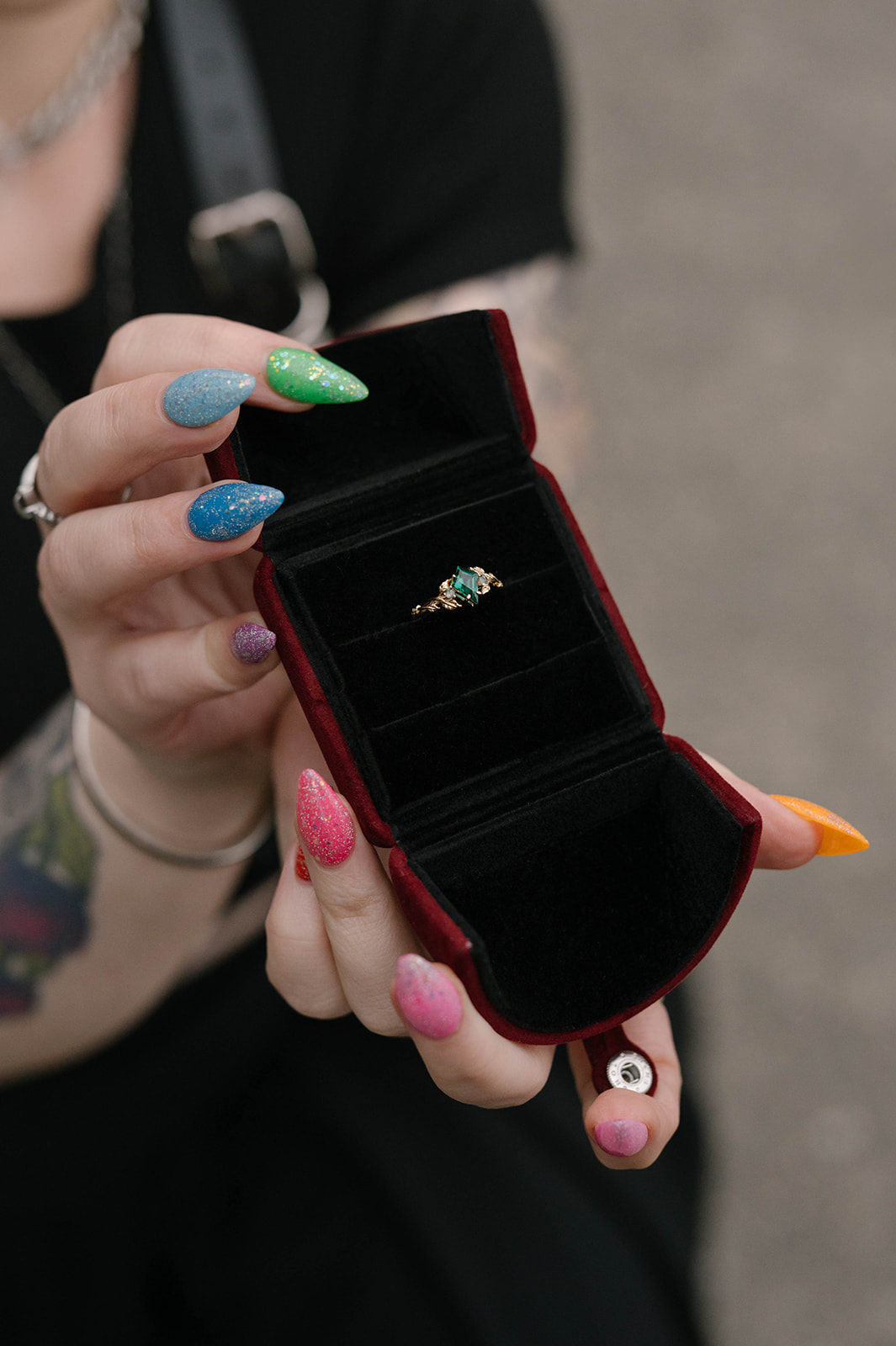 Close-up of pride-colored nails holding an emerald engagement ring in a velvet box.