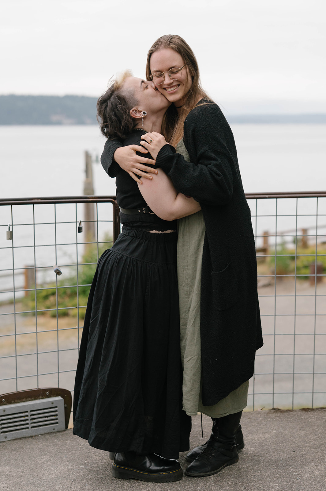 One woman kisses her partner on the cheek as they laugh and embrace at a Whidbey Island overlook above Puget Sound.