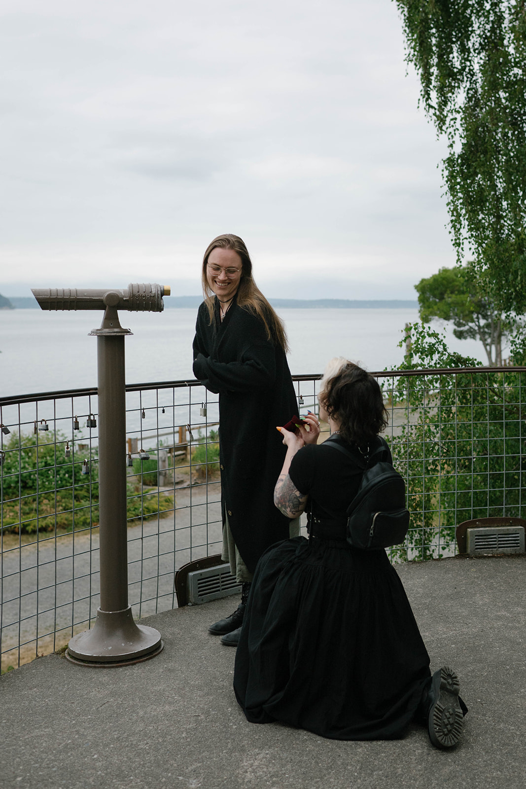 A woman kneels with a ring box while proposing at a whale lookout on Whidbey Island, with the water in the background.