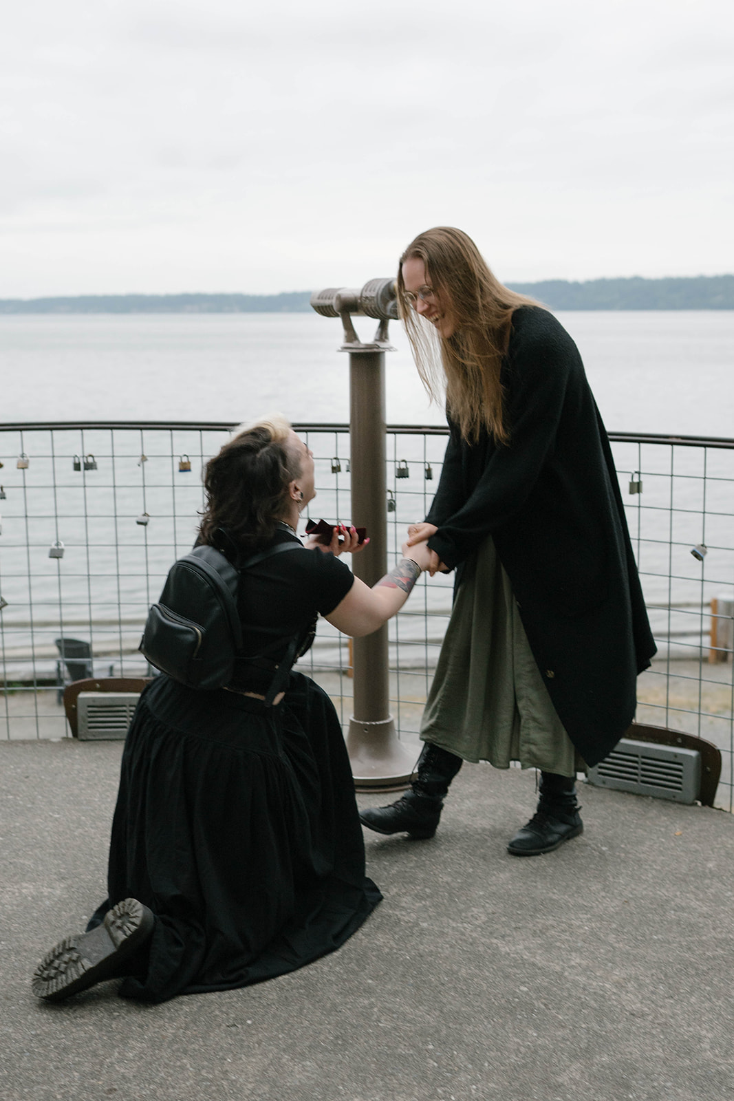 A woman kneels and opens a ring box while proposing at a whale lookout on Whidbey Island, with the water and binoculars behind them.