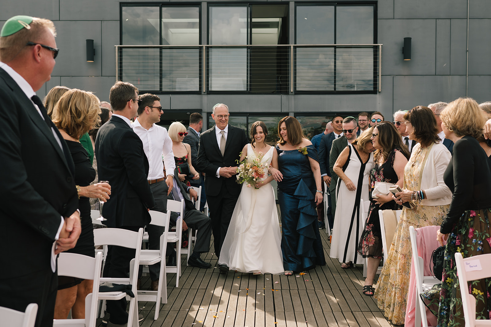 Bride holding her bouquet and smiling as she walks down the aisle with her parents at a Fremont Foundry rooftop wedding