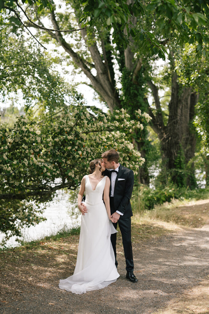 Bride and groom standing under leafy trees beside the water during their Fremont Foundry wedding