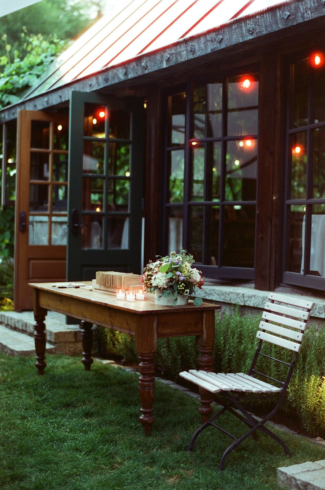 A candlelit wooden table outside the cottage at Bella Luna Farms, surrounded by greenery and warm window light, photographed on Candido ISO 800 film.
