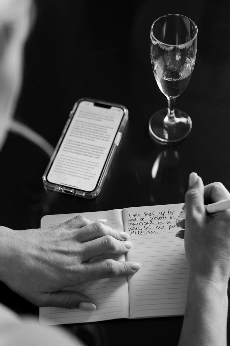 Black and white close-up of the groom writing his vows in a notebook with a glass of champagne and phone beside him.
