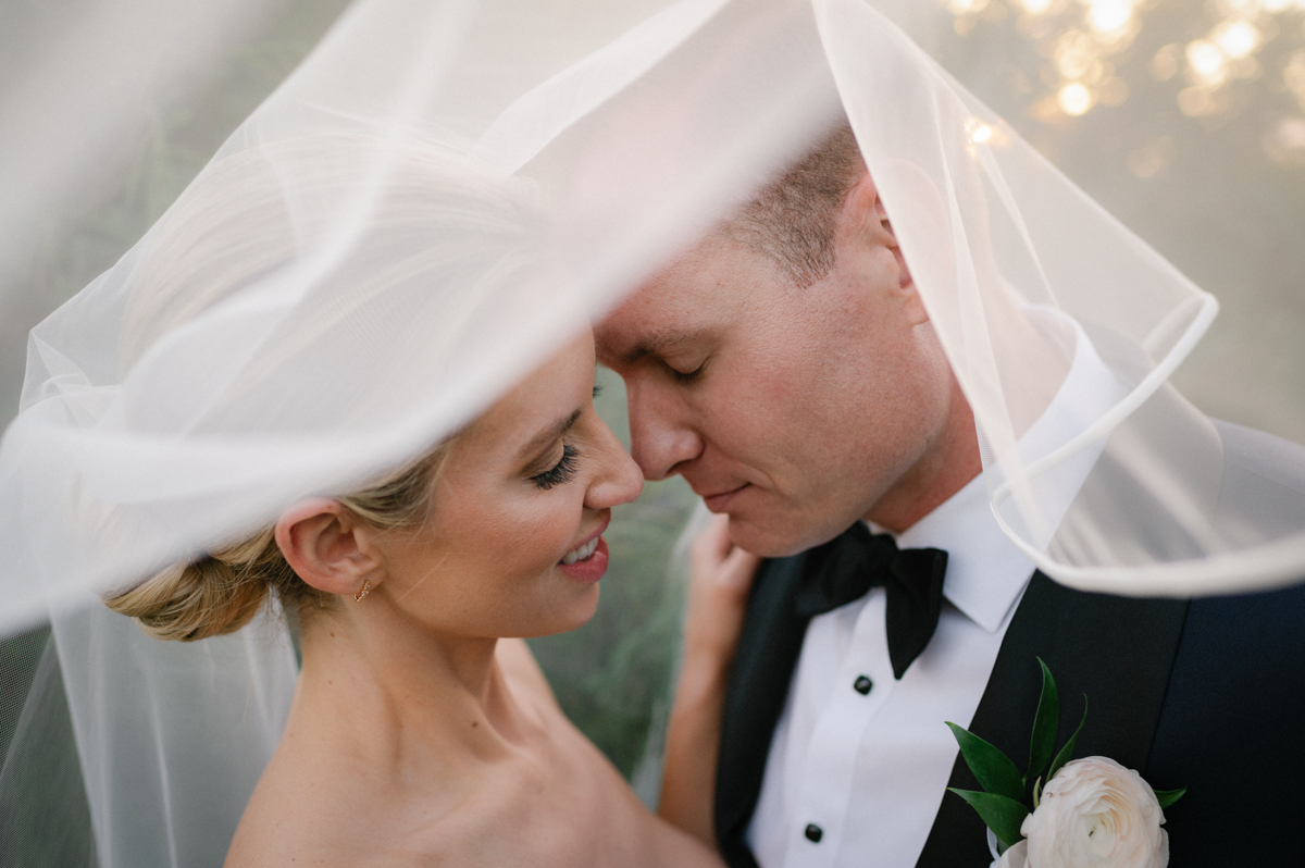 Bride and groom standing close as the bride’s veil lifts in the ocean breeze during golden hour in Kapalua.
