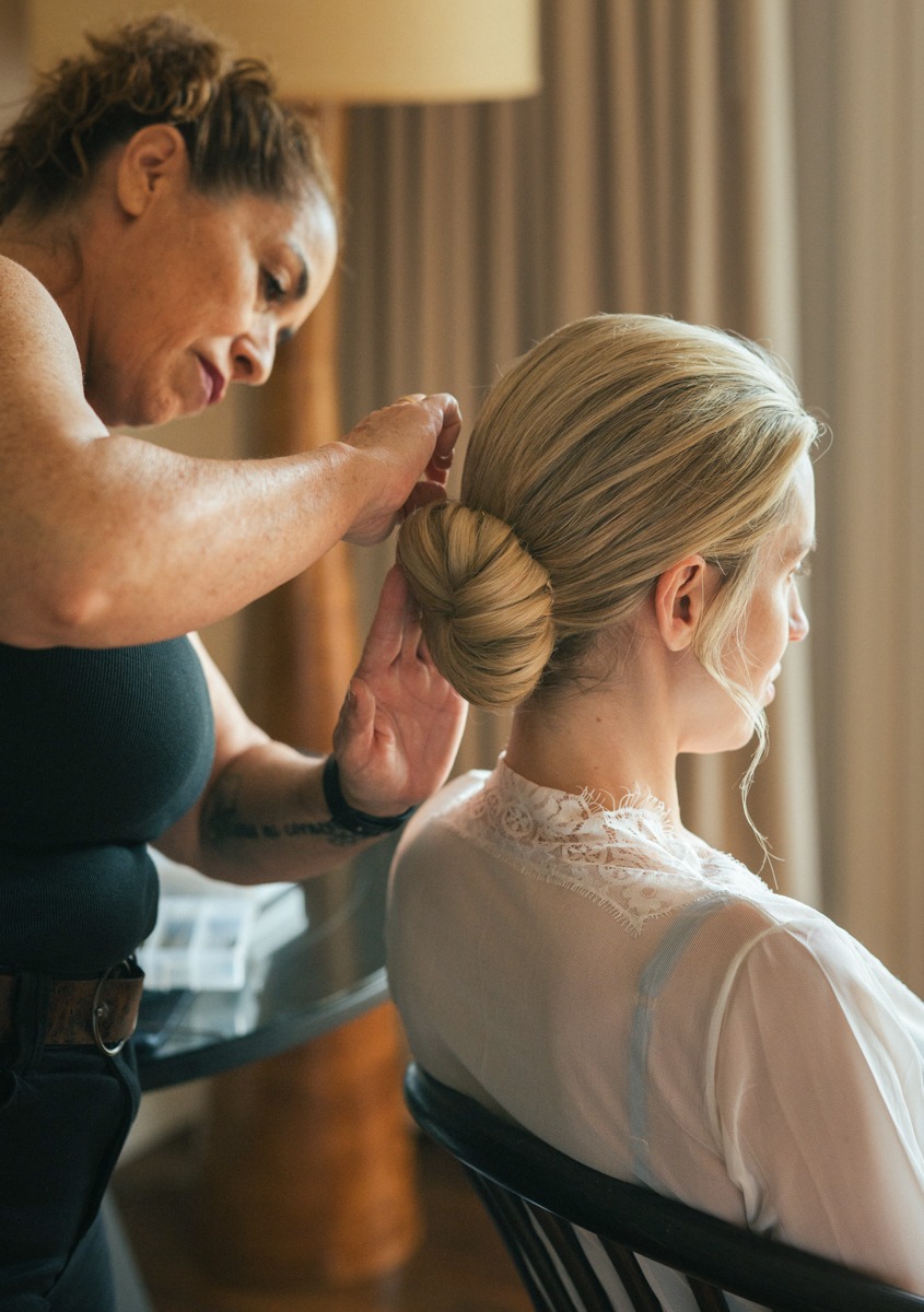 Hairstylist securing the bride’s low bun as she sits in a chair in her lace robe.
