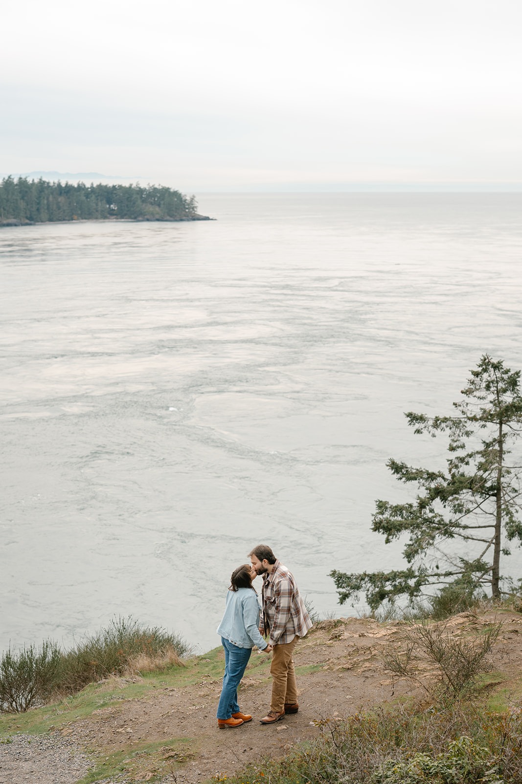 Jonathan and Ashley stand together on a cliff kissing by the water at Deception Pass, small against the vast landscape.