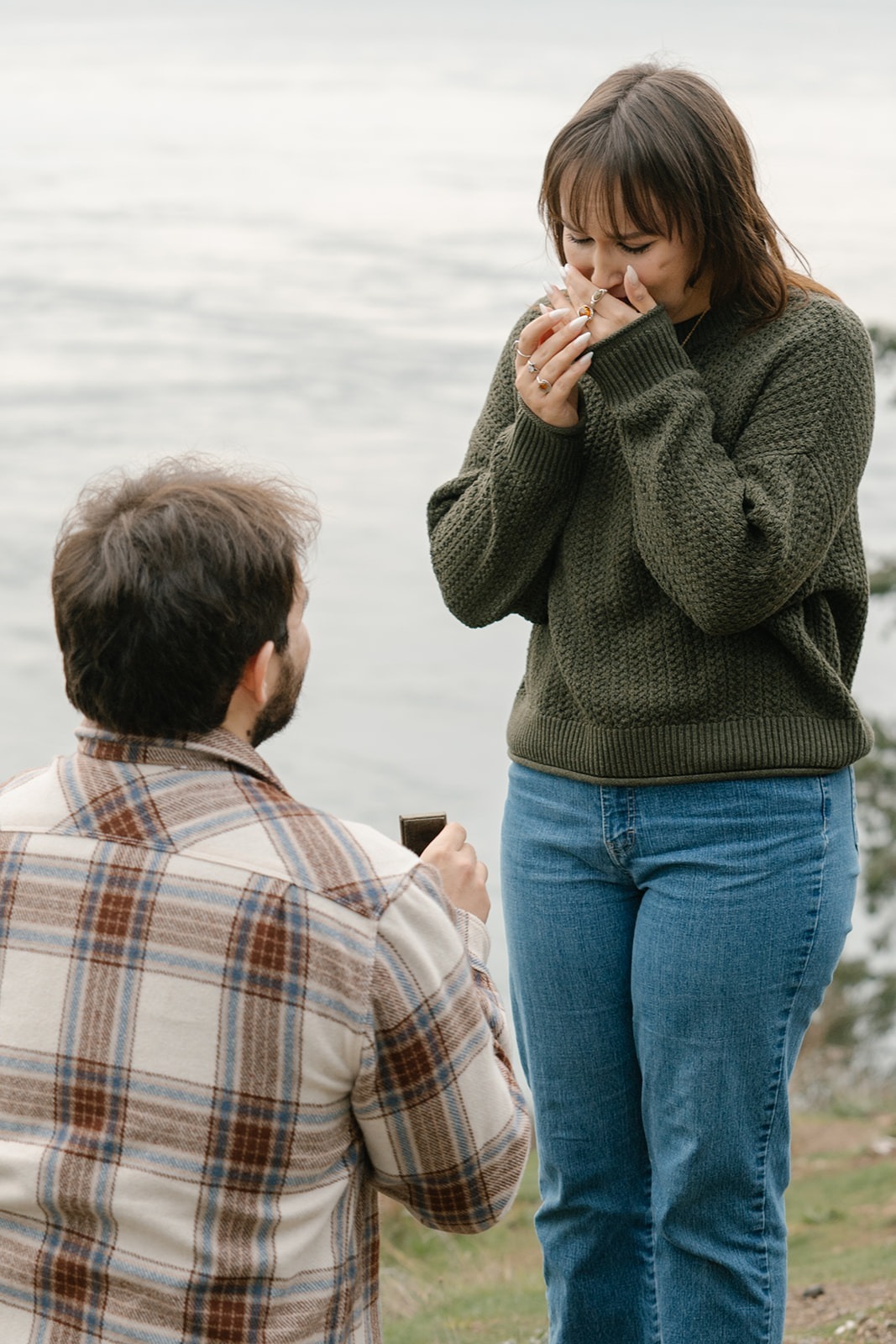 Jonathan kneels in front of Ashley holding the ring box as she stands in stunned happiness.