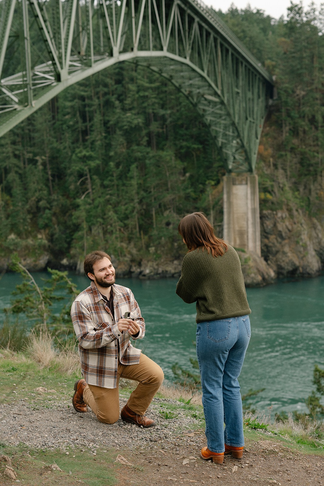 Jonathan gets down on one knee to propose to Ashley with the Deception Pass bridge towering behind them.