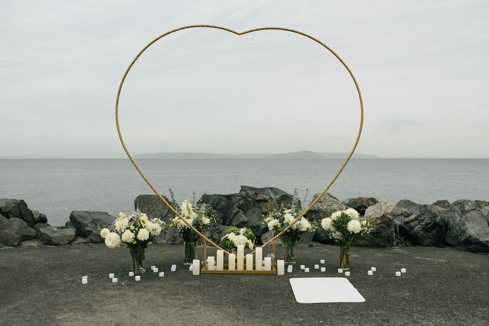 The proposal site prepared with a heart arch, candles, and florals along a rocky waterfront.