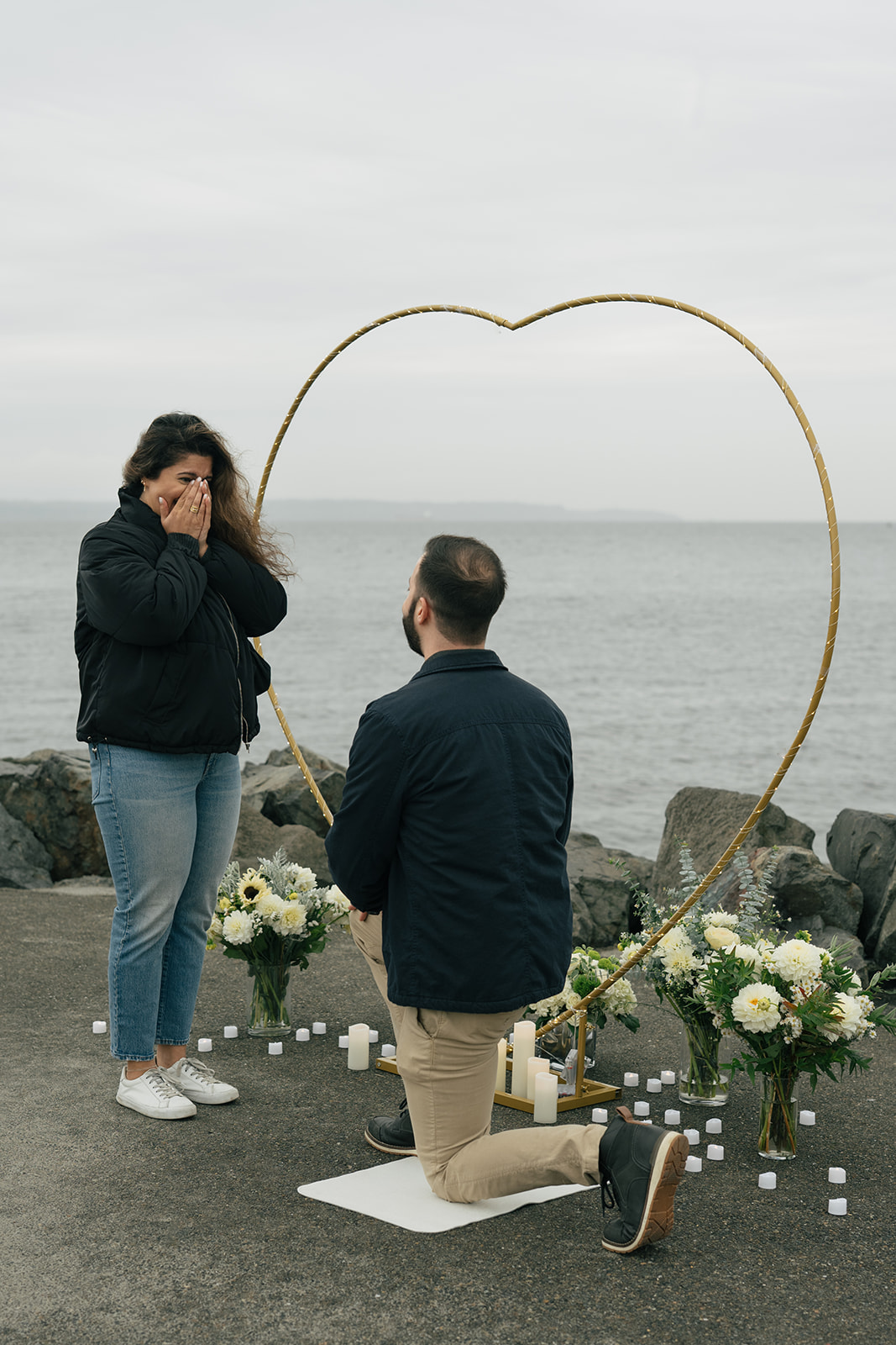 Brad kneels in front of Aysha beneath a heart-shaped arch during their waterfront proposal.