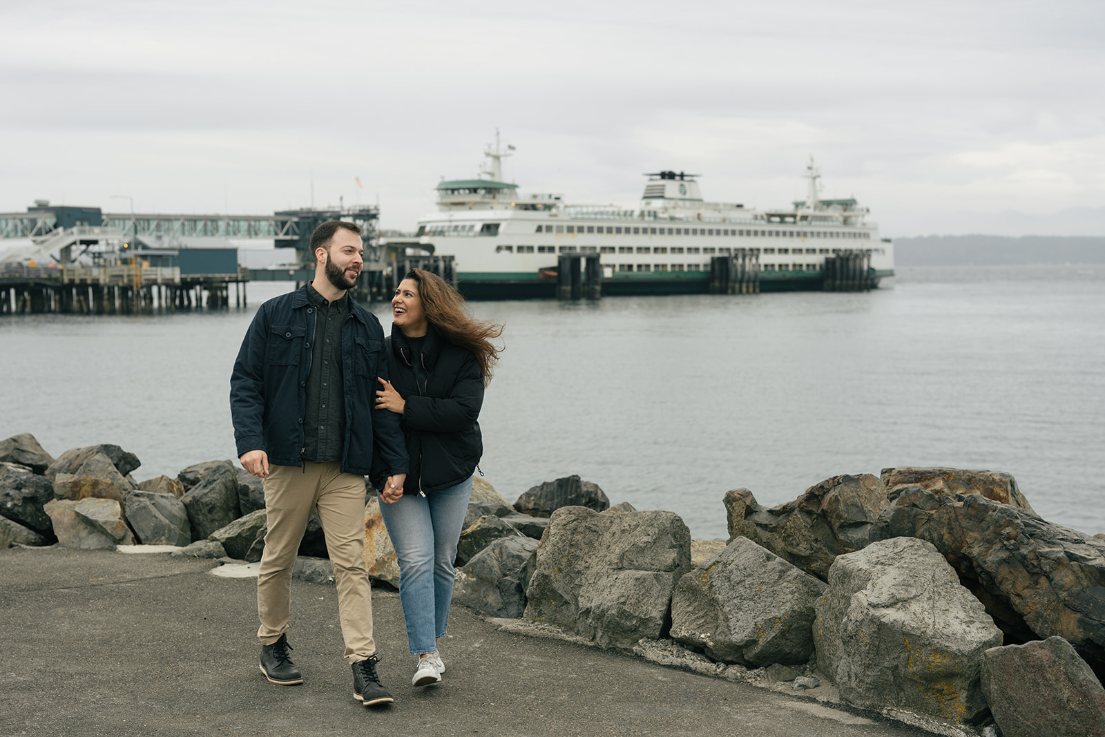Brad and Aysha walk together along the rocky shoreline with a ferry in the background after their proposal.
