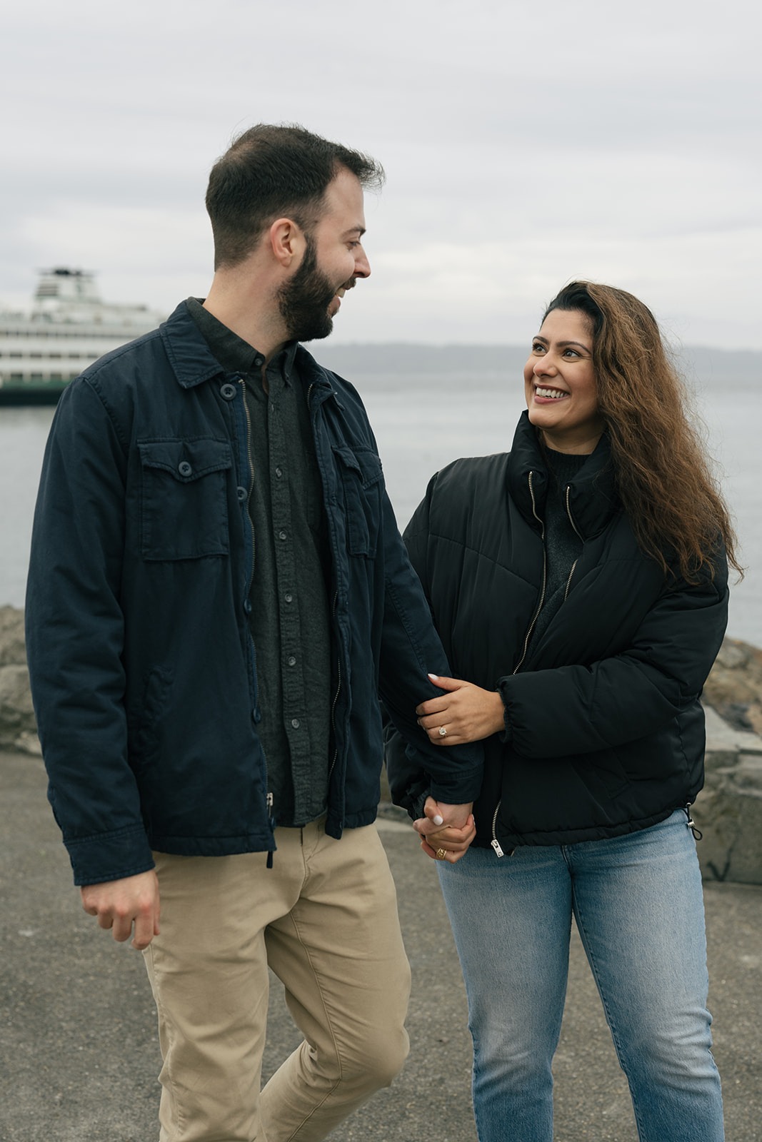 Brad and Aysha smile at each other while walking along the waterfront after getting engaged.