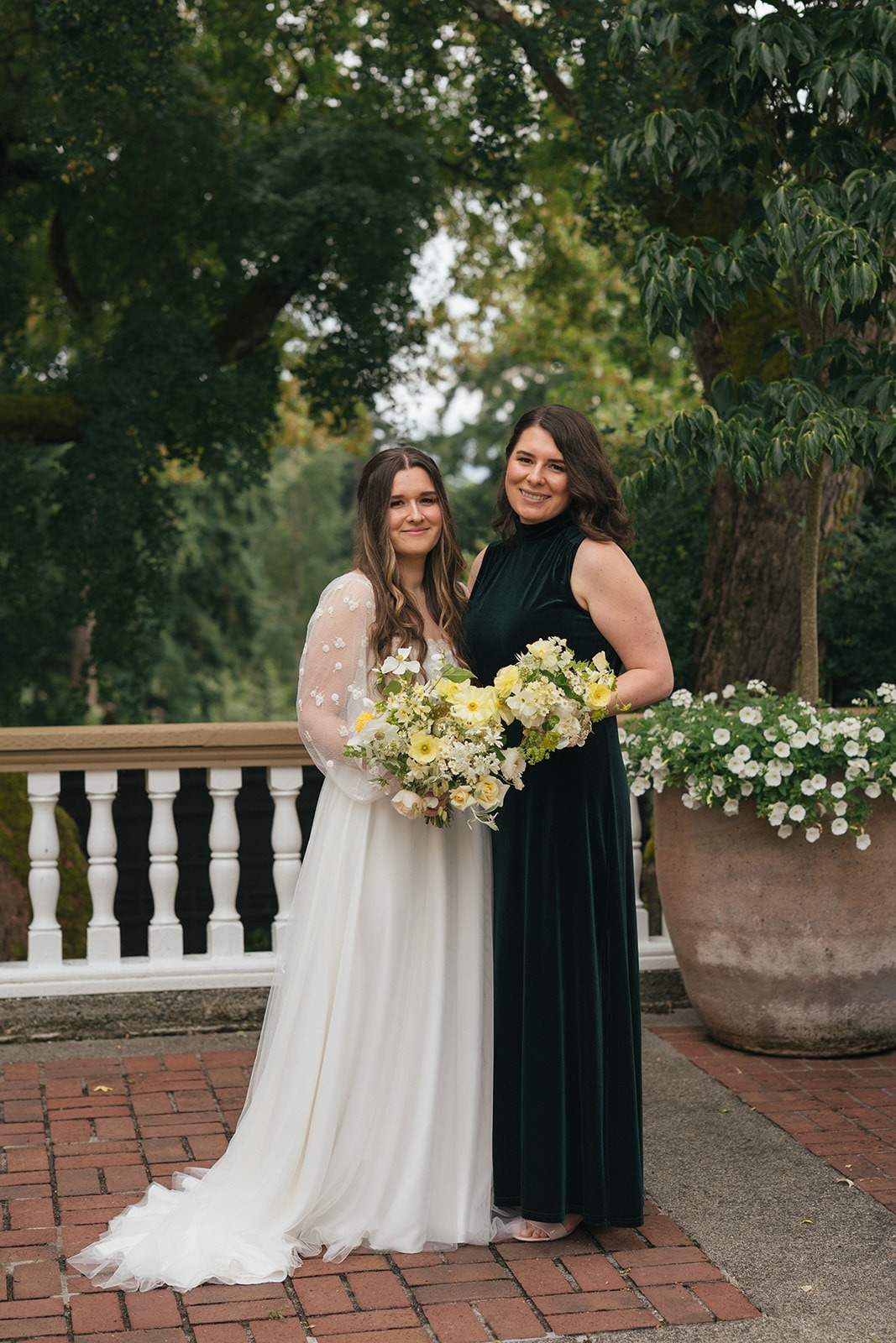 Bride standing with her maid of honor while holding yellow and white bouquets.